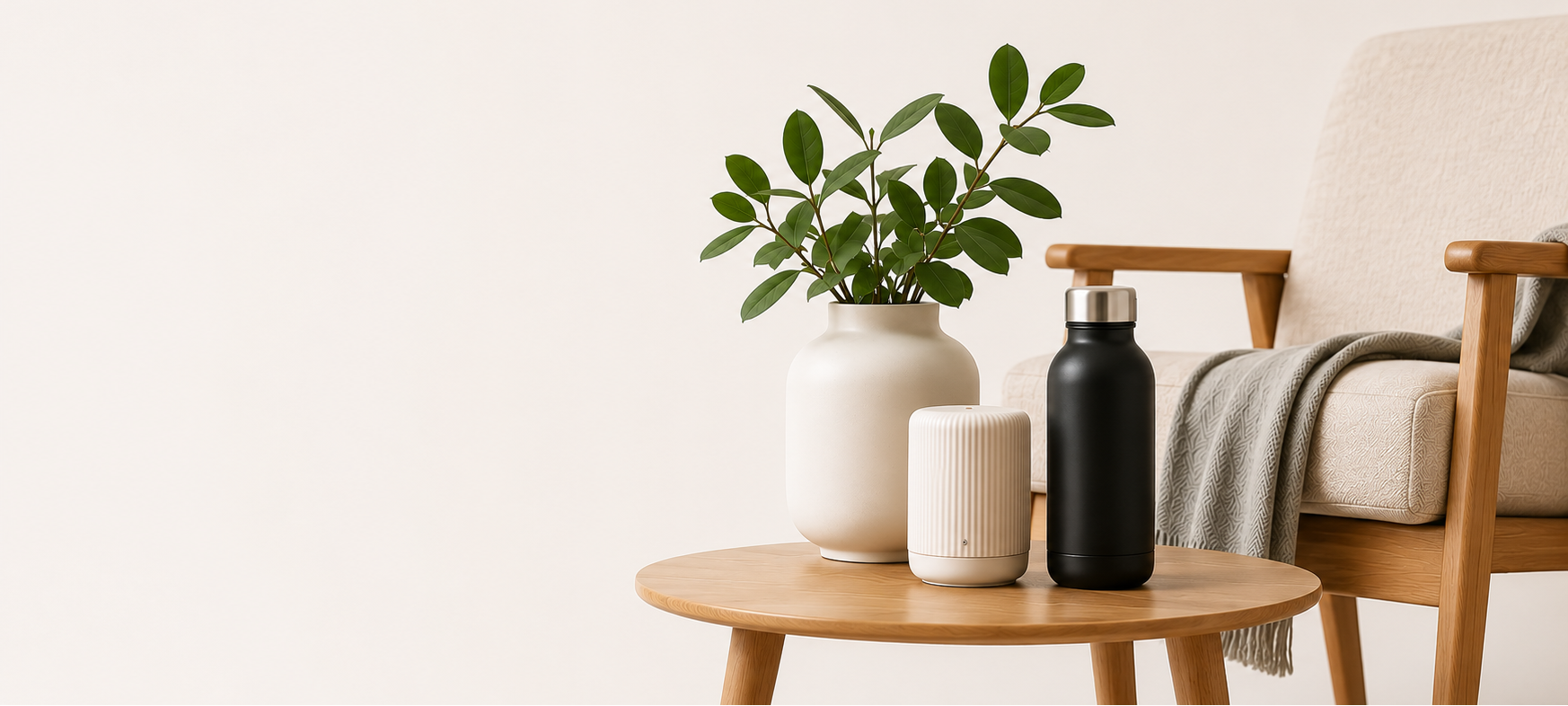 A minimalist interior scene with a small round wooden side table holding a white vase with green leaves, a white cylindrical device, and a black bottle, next to a light upholstered armchair with a gray throw blanket.